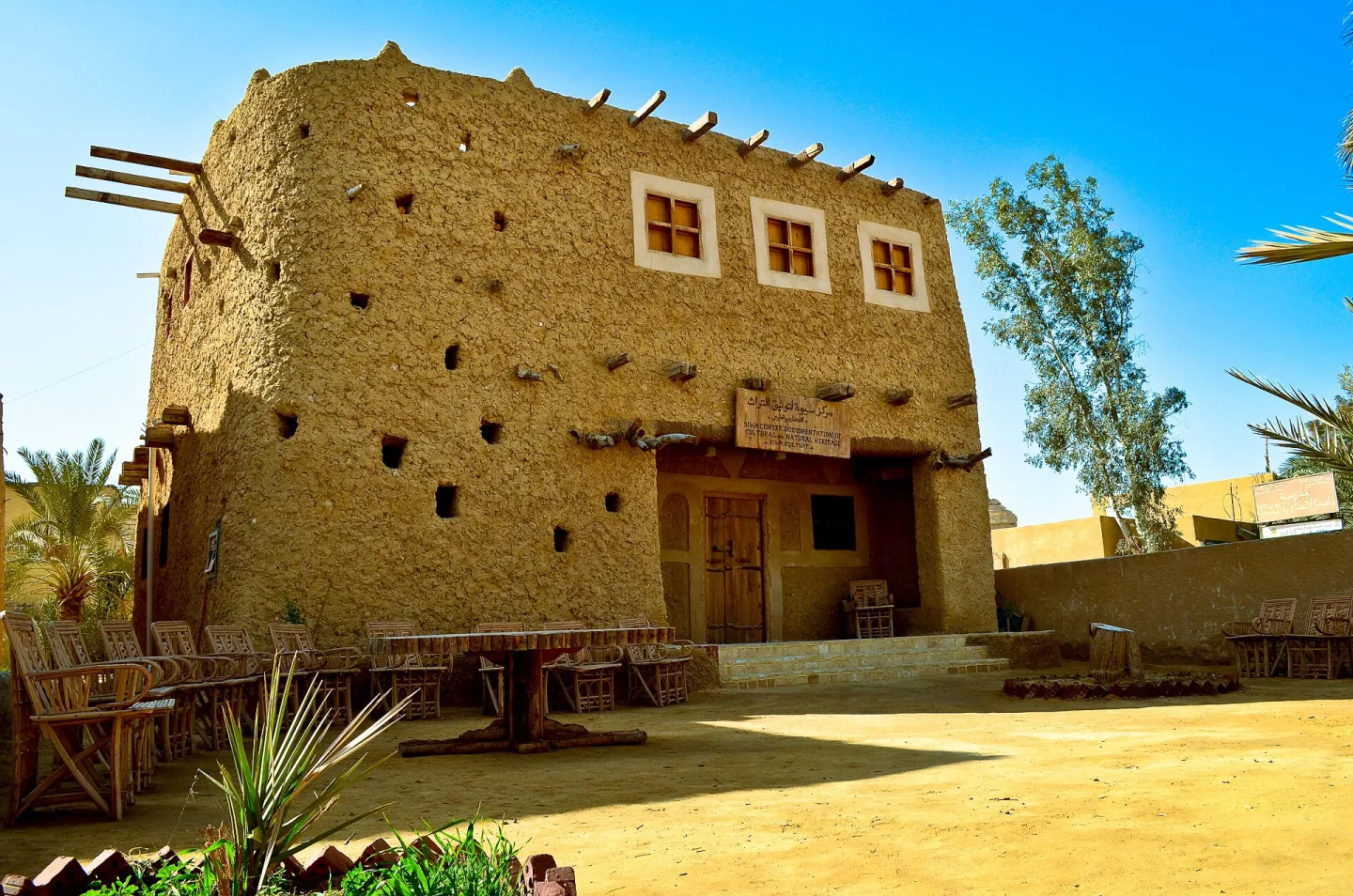 Earth-built Siwa Centre of Documentation building with chairs, palms and a tree